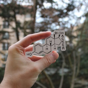 Photo d'un stickers. Il représente le dessin scientifique de la molécule d'œstradiol. En haut à droite, les lettres O et H qui font partie du schéma sont utilisées à la manière d'un mot croisé pour écrire le mot "Biohacker".