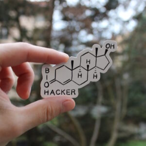 Photo d'un stickers. Il représente le dessin scientifique de la molécule de testostérone. En bas à gauche, la lettre O qui fait partie du schéma est utilisée à la manière d'un mot croisé pour écrire le mot "Biohacker"