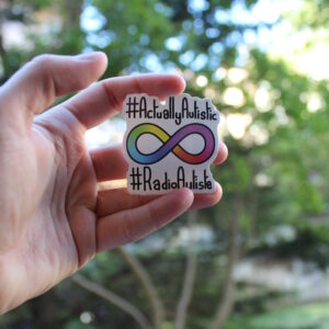 Photo du stickers. Au centre, le symbole de la neurodiversité, un ruban de Moebius multicolore. Au-dessus est écrit en noir "#ActuallyAutistic" et en dessous "#RadioAutiste".