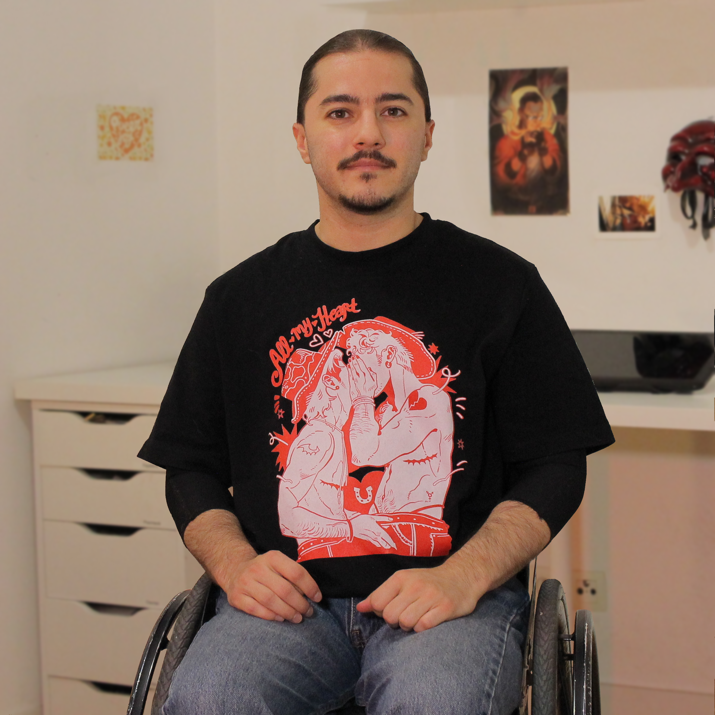 Photo portrait. Alistair est un homme blanc avec les cheveux bruns et longs attachés, une moustache et un bouc. Il est assis dans un fauteuil roulant manuel devant son bureau. Il porte un jean bleu large et un T-shirt noir orné d'un dessins dans les tons rouge et roses de deux cowboys trans qui s'embrassent. 